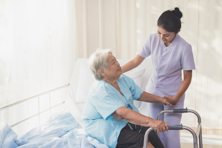 woman caregiver and elderly patient on examination couch. Happy nurse holding hand of senior to help senior patient. female nurse supporting senior disabled woman with walker at hospital. Health care and people concept